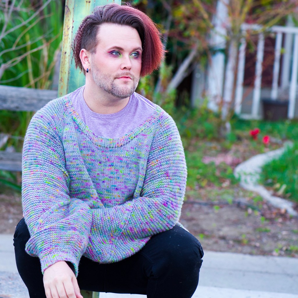 image description: a bearded man with crimped auburn hair squats in front of a lamp post. He is wearing black skinny jeans and an oversized sweater in grey yarn with pops of pink, purple, green, and blue. He also is wearing fabulous eye makeup in shades of purple and pink, with a cut crease in acid green.